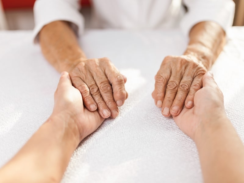 Close-up of carer support dementia with hands of elderly person held gently by caregiver across a white table.