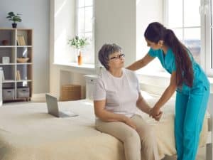 Carer support dementia concept showing a young female caregiver helping an elderly woman sit on a bed at home.