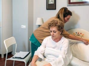 Elderly woman receiving 24-hour personal care as caregiver assists with bed arrangement in a comfortable home setting.