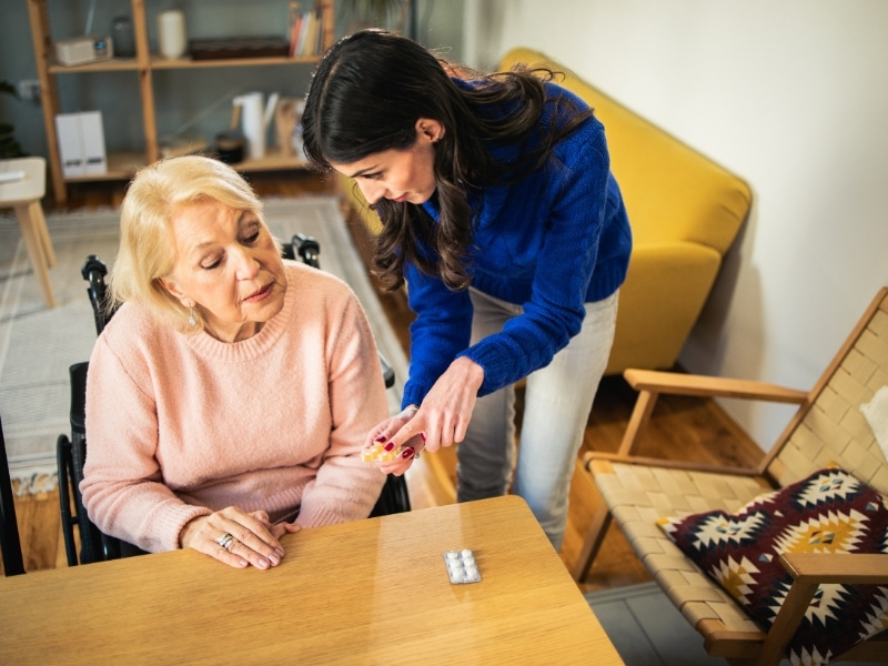 Elderly support with medication assistance—young woman helps senior in wheelchair organise daily pills at home table