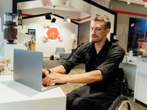 Man in a wheelchair using a laptop at a cafe, showing independence through assistance with travel transport arrangements