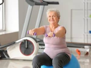 Elderly support for physical health—older woman exercising with hand weights on fitness ball at gym
