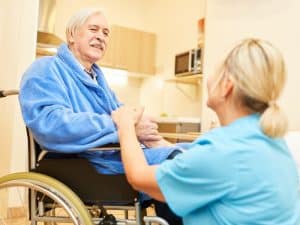 Elderly man in a wheelchair smiling at a nurse in blue uniform, representing home care comfort in a supportive setting