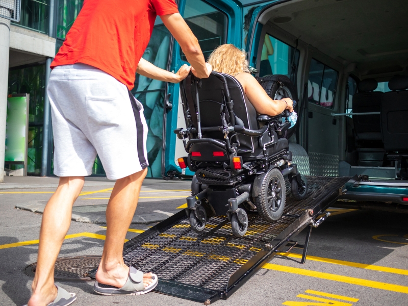 Carer assists woman in wheelchair up a van ramp as part of reliable transportation services for NDIS participants.