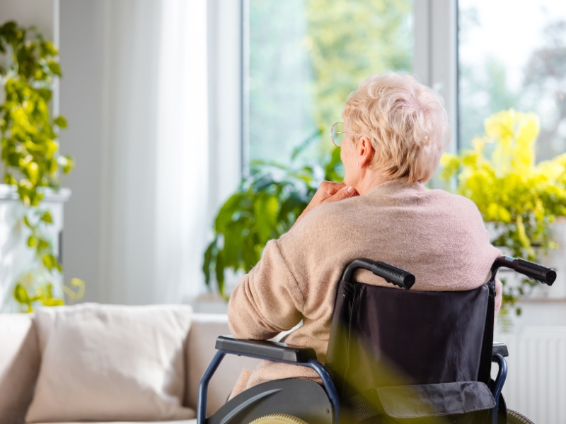 Elderly woman in a wheelchair sits indoors by a window, impacted by lack of reliable transportation services.