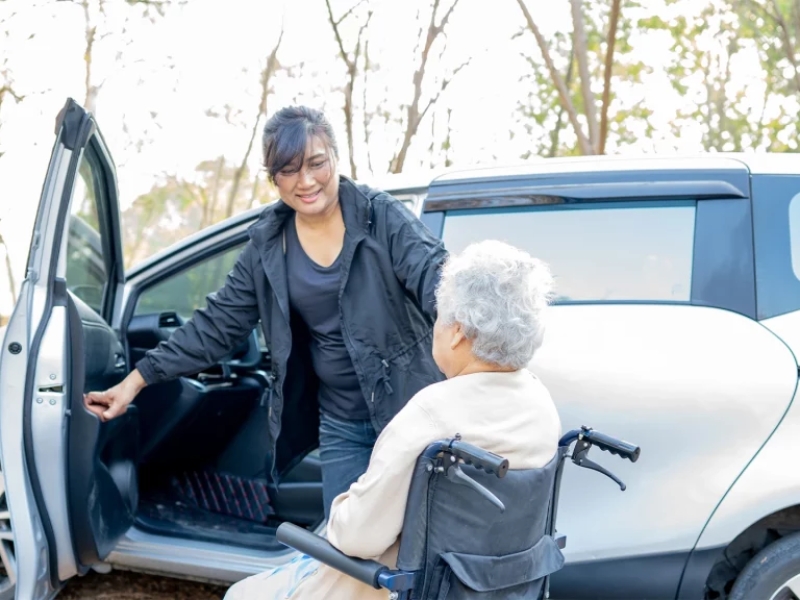 Elderly woman in wheelchair provided with assistance with transportation by caregiver.